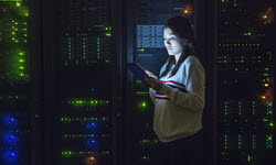 A photograph of a lady looking at a laptop screen in a dark room with other screens in the background