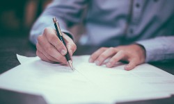 A photograph of someone signing paperwork with a pen in one hand and the other hand on the paper